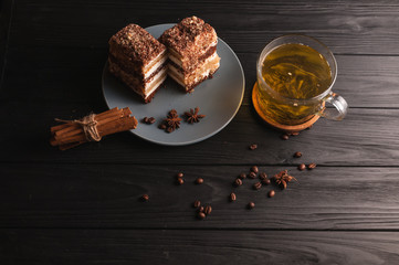 Two pieces of multi-layered honey cake on a plate, on a black wooden background with a cup of tea, chopsticks and coffee grains, top view