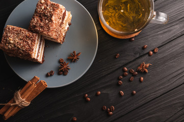 Two pieces of multi-layered honey cake on a plate, on a black wooden background with a cup of tea, chopsticks and coffee grains, top view