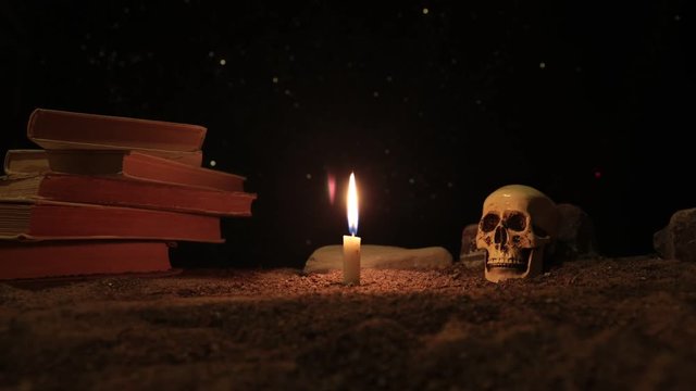 Wizard's Desk. A desk lit by candle light. A human skull, old books on sand surface. Halloween still-life background with a different elements on dark toned foggy background. Slider shot