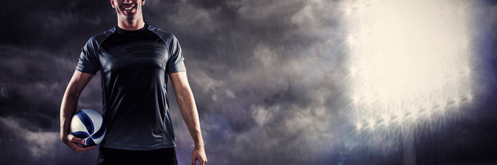 Full length portrait of smiling rugby player in black jersey holding ball against spotlight in the sky