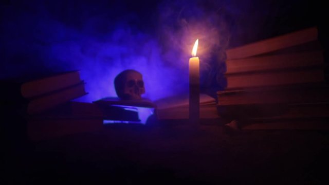 Wizard's Desk. A desk lit by candle light. A human skull, old books on sand surface. Halloween still-life background with a different elements on dark toned foggy background. Slider shot
