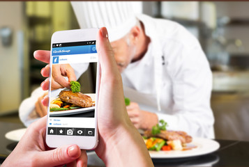 Hand holding smartphone against concentrated male chef garnishing food in kitchen