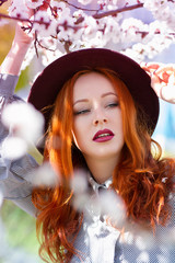 Stylish young beautiful red-haired girl in a burgundy hat near a flowering tree - 203176067
