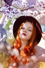 Stylish young beautiful red-haired girl in a burgundy hat near a flowering tree - 203176094