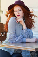 Stylish young beautiful red-haired girl in a burgundy hat in a street cafe - 203176234