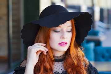 Stylish young beautiful red-haired girl in a black hat in a street cafe - 203176274