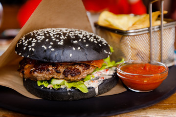 Close-up of black burger with sesame, juicy cutlet, french fries on paper backing.