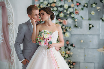 beautiful wedding couple hugging in bright studio. The groom in a business gray suit, a white shirt in a bow tie and a buttonhole, a bride in a luxury dress with a veil and a bouquet of flowers