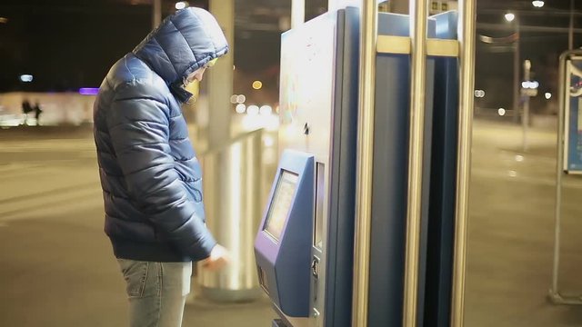 A young man in a warm jacket goes to the parking machine on the street in the city.