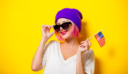 Young girl with pink hair in purple hat and sunglasses holding United States flag on yellow...