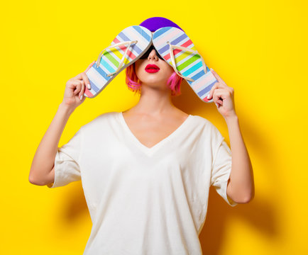 Young girl with pink hair in purple hat and sunglasses holding a flip flops shoes on yellow background