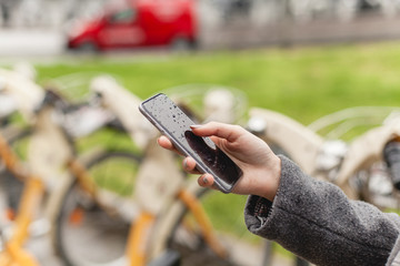 Closeup shot of a young girl using bicycle rent mobile app outdoors, hands of female manager browsing smartphone standing near bike sharing to go cycling, concept of healthy lifestyle in the city