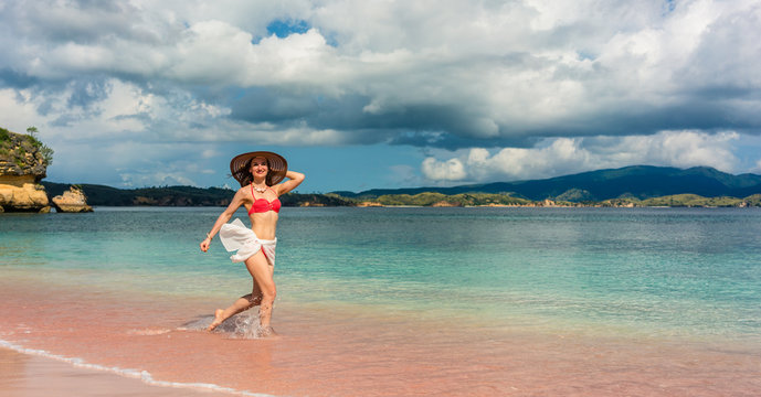 Full length of a happy young woman wearing straw hat and a trendy swimwear while walking through shallow sea water on the beach in Komodo Island, Indonesia