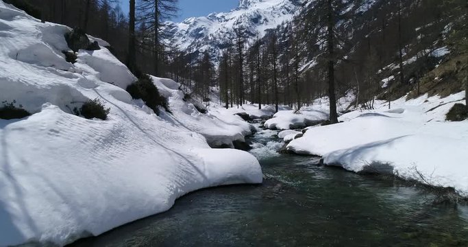 Flying over stream in snowy valley on a sunny spring day. Europe Italy Alps outdoor wild nature, aerial drone flight.