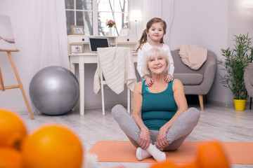 Fototapeta premium Happiness. Cheerful old grandmother sitting on the carpet and her granddaughter hugging her from behind