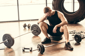 upset young sportsman sitting with dumbbells and barbells in gym