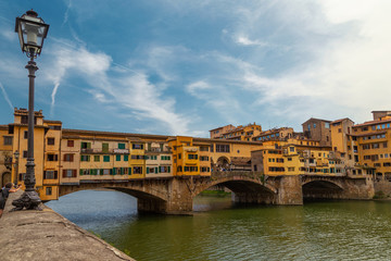 Obraz premium View on The Ponte Vecchio bridge on a sunny day, a medieval stone segmental arch bridge over the Arno River, in Florence, Italy, noted for still having shops built along it