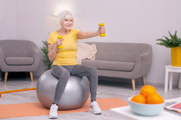 Exercising cheerfully. Joyful elderly woman posing for the camera while sitting on the balance ball and working out with dumbbells