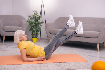Strengthening exercise. Charming elderly woman sitting on the yoga mat and doing leg lift exercise...