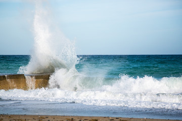 Wasserkraft. Eine Welle klatscht mit voller Wucht gegen einen Felsen an der Küste Kretas, im Vordergrund Sandstrand und Brandung, im Hintergrund das Mittelmeer und blauer Himmel .