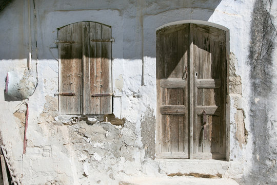 Schlicht und dennoch sch&ouml;n, eine durch Regen und Sonne verwitterte, robuste und antike Holzt&uuml;r mit Fensterl&auml;den aus Holz, umgeben von einer abbr&ouml;ckelnden und verwitterten weiss gekalkten Hausfassade. 