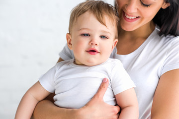 partial view of smiling mother with cute little baby in hands at home