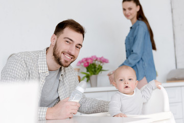 father feeding son with milk in baby bottle, mother standing on kitchen behind