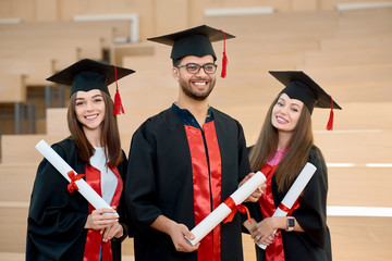 Smiling graduates keeping diplomas standing in spacy modern university classroom. Students wearing new black and red graduation gowns. Feeling happy, satisfied, laughing. Having brilliant future.