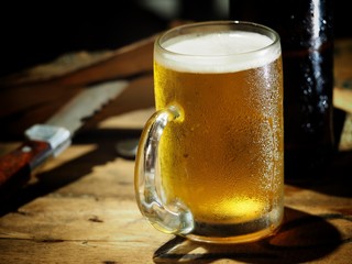 Beer in a glass and a bottle on a wooden table for work, with a knife and a log on the side.