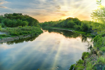 Sunset over river. Landscape with green trees, dramatic sky, clouds, sun and sunrays