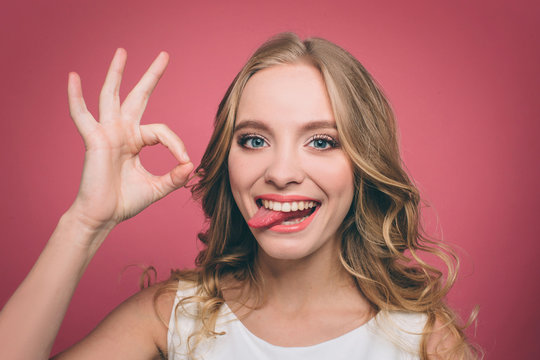 Funny young woman is looking straight forward and showing a piece of her tounge while she is smiling. Also girl is showing the ok symbol with her fingers. Isolated on pink background.