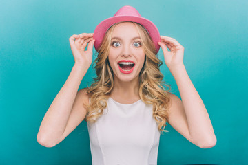 Happy and amazed girl is looking straight on camera. She is very excited. Girl is holding hat with her hands. Isolated on blue background.