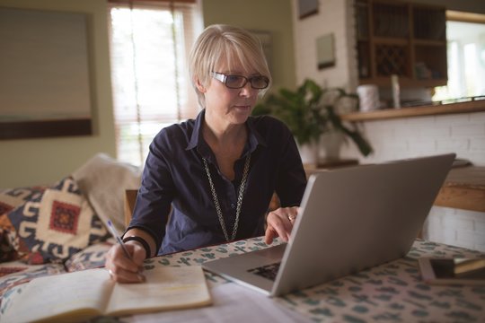 Mature woman using laptop while writing in notebook