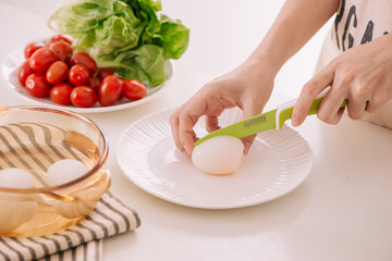 Close up of females hands cutting boiled egg for his breakfast and mixed vegetables on white wooden background