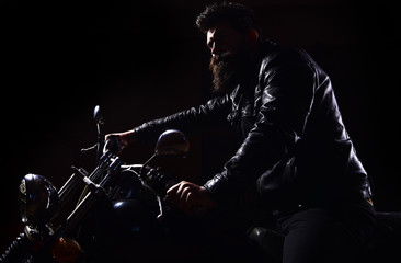 Man with beard, biker in leather jacket sitting on motor bike in darkness, black background. Night...