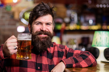 Guy spend leisure in bar, defocused background. Man on happy smiling face sit in bar or pub near bar counter. Hipster with beard holds glass with beer, raising up, cheers. Cheers concept.