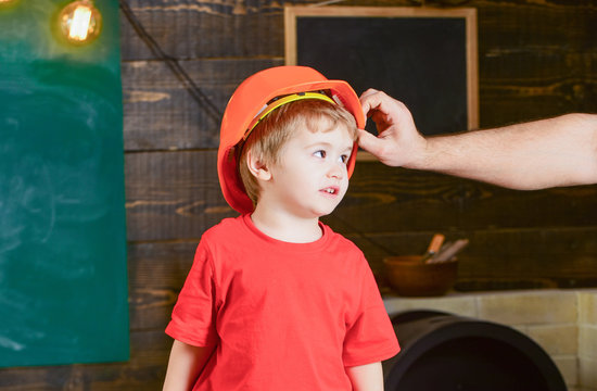 Toddler In Protective Hard Hat, Helmet At Home In Workshop. Child Cute And Adorable Stands While Male Hand Putting Hard Hat On Head. Protection And Safety Concept. Carefully Protect Kid With Helmet.