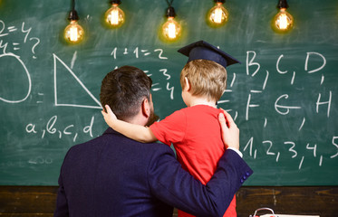 Teacher with beard, father hugs little son in classroom while discussing, chalkboard on background....