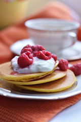 Four corn pancake lie on a white plate with sour cream and raspberry berries, on the background of a Cup of tea.