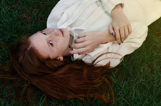 A Beautiful Girl With Long Red Hair And Brown Eyes, Dressed In A White Blouse Lies On The Green Grass.