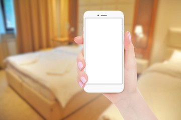 Female hand holding modern white smartphone with isolated screen, hotel bedroom in background. Travel concept, mockup