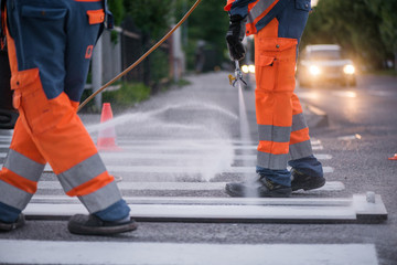 Traffic line painting. Workers are painting white street lines on pedestrian crossing. Road cones with orange and white stripes in background, standing on asphalt during road construction works. 