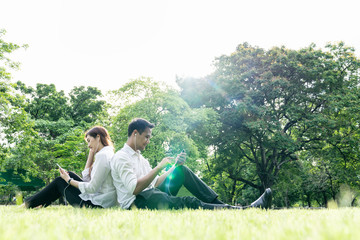 Naklejka na ściany i meble Young asian couple,lover holding earphone,smartphone sit relax in park,tree background copy space. Happy outdoor on summer,spring sunshine in city green park,garden lifestyle. Take a break after work.