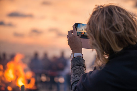 Woman is taking picture of sunset with smart phone. Romantic bonfire night at seaside. People gathering together to celebrate Night of ancient lights. Large burning campfire with soft glowing flame