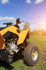 A four-wheeled yellow ATV quad-bike standing idle on the green grass, with trees and a blue sky...