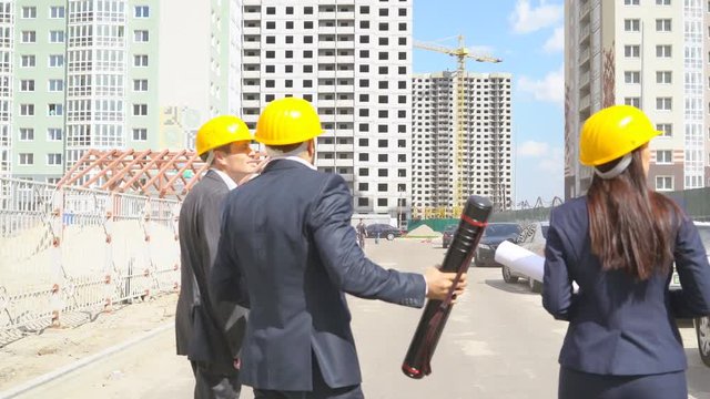 three serious business people on the construction site are on the road. Men and woman in suits and yellow helmets. people on the background of high-rise buildings under construction 4K