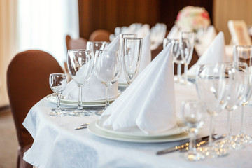 Glasses, flowers, fork, knife, napkin folded in a pyramid, served for dinner in restaurant with cozy interior. Wedding decorations and items for food, arranged by the catering service on a large table