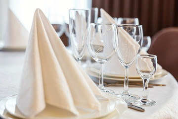 Glasses, flowers, fork, knife, napkin folded in a pyramid, served for dinner in restaurant with cozy interior. Wedding decorations and items for food, arranged by the catering service on a large table