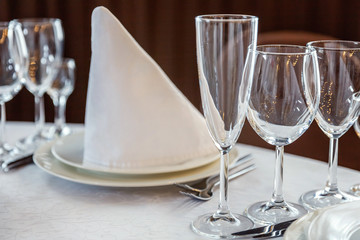 Glasses, flowers, fork, knife, napkin folded in a pyramid, served for dinner in restaurant with cozy interior. Wedding decorations and items for food, arranged by the catering service on a large table