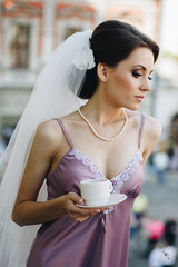 Elegant brunette bride woman in white bridal veil and beautiful nightgown drinking morning coffee on the balcony before the wedding ceremony.
